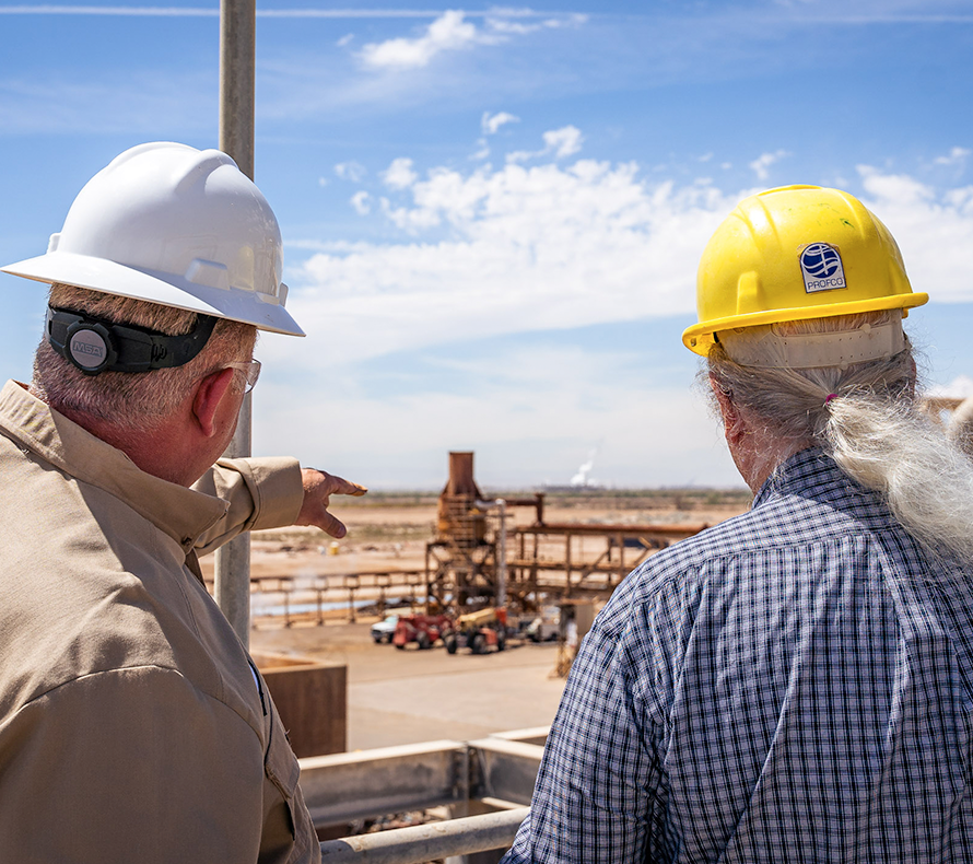 Two men in hard hats look out at a geothermal power plant, with one pointing into the distance