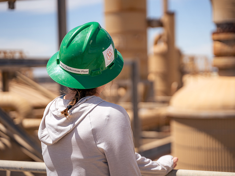 A woman in a hardhat looks out at a complex power plant, which is out-of-focus in the background